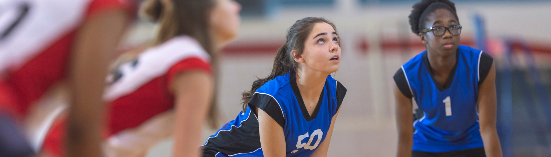 Adult Volleyball League Header | 2 girls wearing volleyball jerseys crouched and waiting for a ball with two teammates out of focus