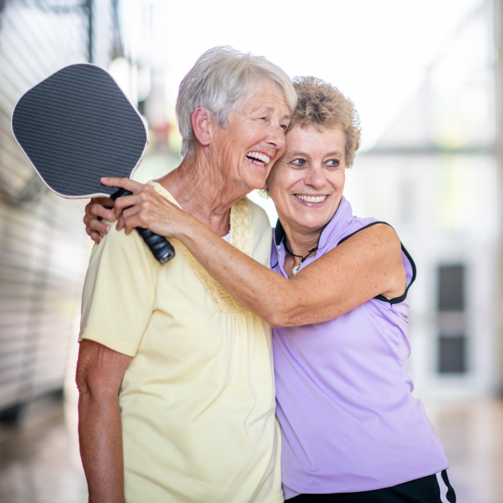 2 older female pickleball players hugging after playing on an indoor pickleball court